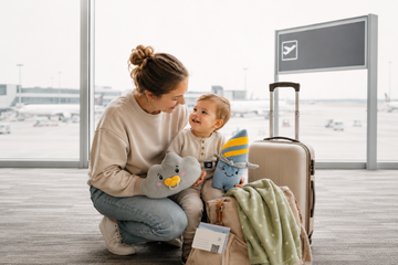 Mutter und Kleinkind am Flughafen mit Reisegepäck und Lenabies als vertraute Begleiter für den ersten Urlaub mit Baby