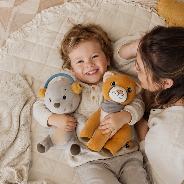 Fröhliches Kleinkind liegt mit Mutter auf weicher Decke und kuschelt mit den Lenabies Aaron dem Bären und Tali der Puma Dame. Interaktive Kuscheltiere für Kinder begleiten gemeinsame Familienmomente in einer warmen und geborgenen Atmosphäre.